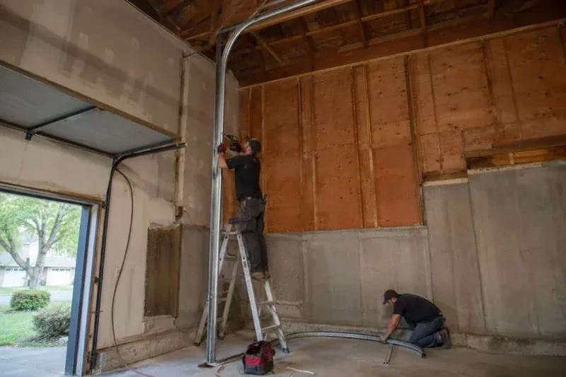 One person on a stepladder working beside a tall curved garage-door track while another kneels beside loose track sections in an unfinished garage. — High-Lift Garage Door Conversion, Oshawa